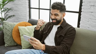 A cheerful young man with a beard celebrates a victory at home while holding a smartphone clipart