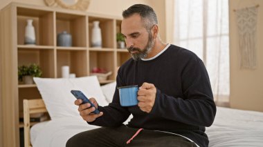 Beyaz sakallı, akıllı telefon kullanan ve elinde kahve fincanı tutan olgun bir İspanyol.