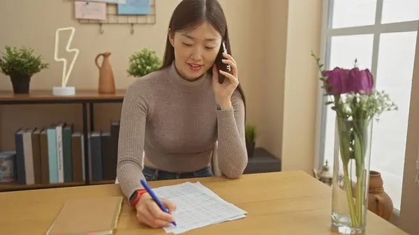 stock image A young asian woman talks on the phone while writing notes at a wooden table in a well-lit home interior.