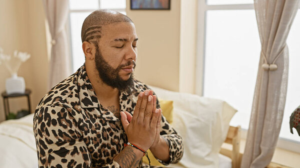 Африканский американец молится в спокойной спальне с скрепленными руками, с закрытыми глазами и мирным выражением.