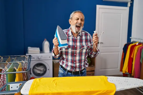 stock image Elderly man's hearty laughter fills the laundry room, portrait of a cheerful senior, ironing chores made fun by a hilarious joke!