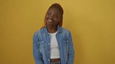 Kot ceket giyen gülümseyen genç bir Afrikalı kadın izole edilmiş sarı bir arka plana karşı duruyor.