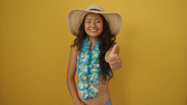 Şapkalı ve çiçekli bir çelenk takan genç Çinli kadın sarı arka plana karşı baş parmak işareti yapıyor.