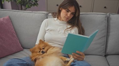 Bir kadın, rahat bir oturma odasında kanepede Pomeranian köpeğiyle kitap okuyarak rahatlıyor..