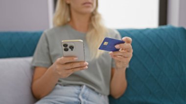 Evde kredi kartı ve akıllı telefon tutan genç sarışın bir kadın çevrimiçi alışveriş veya evde bankacılık tasvir ediyor..