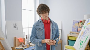 Kıvırcık saçlı ve gözlüklü modaya uygun genç bir adam bir sanat stüdyosunda elinde akıllı bir telefonla dikiliyor..