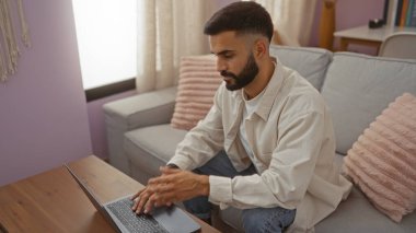Sakallı genç İspanyol bir adam rahat bir oturma odasında kanepe, yastık ve hafif ışıklandırmayla dizüstü bilgisayarla çalışıyor..