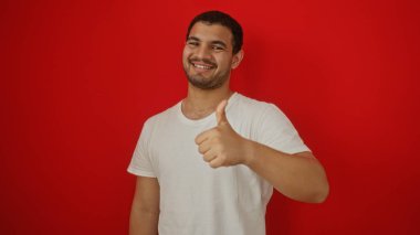 Genç İspanyol bir adam, izole edilmiş kırmızı bir arka planın önünde, beyaz bir gömlek giyiyor ve özgüvenle gülümsüyor.