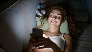 Genç, beyaz bir kadın karanlıkta akıllı telefonuna bakarken yatakta gülümsüyor..