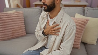 Oturma odasında eli göğsünde oturan genç, İspanyol bir adam, yastıklarla ve ev dekoruyla çevrili, modern, rahat bir dairede rahatsızlık veya acı hissi veriyor..