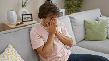 Upset young man sitting on a gray couch at home, covering his face with his hands in a gesture of despair clipart