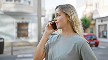Sarışın bir kadın, güneşli bir caddede yürürken telefon eder.