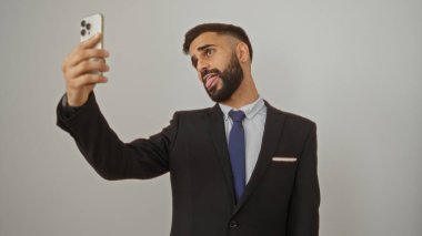 İspanyol genç adam, izole edilmiş beyaz arka plan duvarına karşı takım elbise giyerken akıllı telefonuyla komik bir selfie çekiyor..