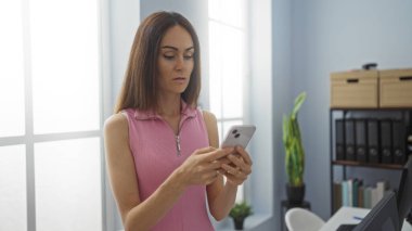 Ofiste pencerenin yanında dururken akıllı telefon kullanan genç bir kadın.