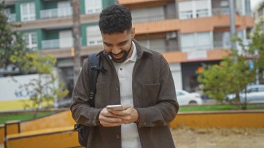 Sakallı, gülümseyen genç bir İspanyol, şehir parkında akıllı telefon kullanıyor, rahat, yaklaşılabilir bir stil sergiliyor..
