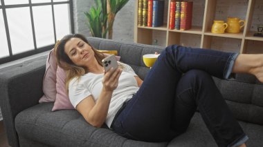 Rahat İspanyol bir kadın, rahat bir oturma odasında elinde telefon ve kahve bardağıyla kanepede uzanıyor..