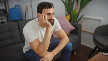 Düşünceli sakallı bir adam, gündelik giysiler içinde, orta karar dekore edilmiş bir oturma odasında, rahat bir koltukta oturuyor..