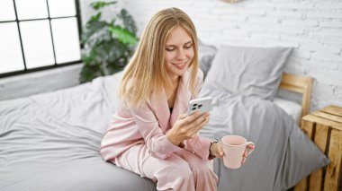 Pijamalı sarışın kadın akıllı telefon kullanıyor ve parlak yatak odasında kahve fincanı tutuyor.