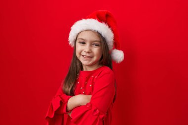 Sevimli İspanyol kız, kollarını kavuşturmuş, kırmızı arka planda noel şapkası takıyor. Onun mutlu, ışıltılı gülümsemesi kameraya ışıldıyor. Ne kadar iyimsersin.!