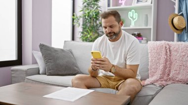 Normal giyimli, olgun sakallı bir adam. Kapalı bir koltukta akıllı telefon kullanıyor. Evde boş vaktini somutlaştırıyor..