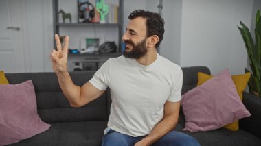 Beyaz gömlekli sakallı adam modern oturma odasındaki kanepede otururken barış işareti yapıyor..