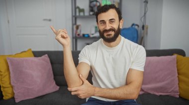 Sakallı bir adam, gri bir koltukta renkli yastıklarla oturma odasında el kol hareketi yapıyor..