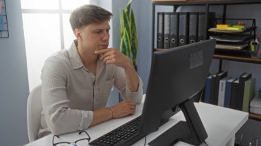 Ofisteki genç adam arkaplanda raflar ve dosyalar olan bir bilgisayar ekranına bakarken düşünüyor.