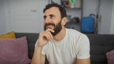 Sakallı, düşünceli, orta yaşlı bir adam modern bir salonda oturuyor. Kendine güveni tam ve sıradan bir tarzı var..