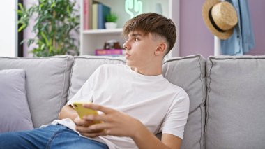 Kafkasyalı genç bir beyaz çocuk elinde akıllı bir telefonla bir kanepede uzanır. Oturma odasının arka planına yaslanır..