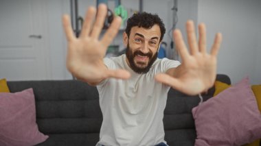 Sakallı bir adam modern bir oturma odasında elleri ile durdurmak, oyuncu görünümlü ve gülümsüyor.