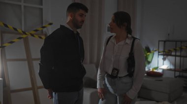 Bir erkek ve bir kadın, muhtemelen dedektifler, loş bir suç mahallinde dururlar..