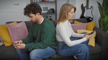 Bir erkek ve kadın, arka planda renkli yastıklar ve gitarla modern bir oturma odasında arka arkaya akıllı telefonları kullanarak koltukta sırt sırta otururlar..