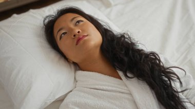 Genç Çinli bir kadın rahat bir yatak odasında bir yatakta dinleniyor, güzel uzun saçlarını ve ev ortamında huzurlu ifadesini sergiliyor.
