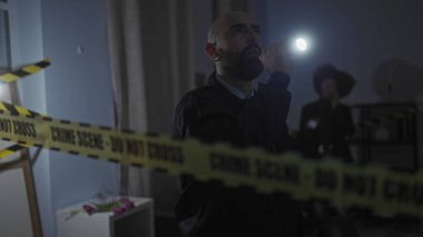 Man and woman investigators with flashlights search a dimly lit indoor crime scene cordoned with caution tape. clipart