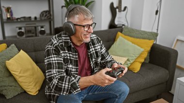 Gülümseyen orta yaşlı bir adam, rahat bir oturma odasında kulaklık takıp kumandayla oynamaktan zevk alır..