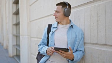 Kulaklıklı, şehir ortamında tablet kullanan, odaklanmış ve sıradan görünen Latin kökenli genç bir adam..