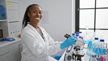 Laboratuvar önlüğü giymiş gülümseyen bir kadın bilim adamı modern bir laboratuvar ortamında mikroskop kullanıyor..