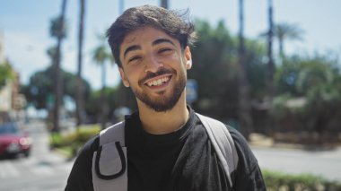 Sırt çantası ve sakallı gülümseyen genç adam palmiye ağaçlarıyla kaplı güneşli bir şehir sokağında duruyor.