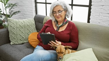 Gri saçlı bir kadın, sıcak bir oturma odasında evcil köpeğiyle birlikte tabletinde kitap okumaktan hoşlanır..