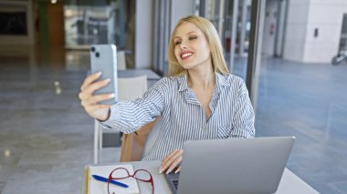 Masasında akıllı telefonu, gözlüğü ve laptopuyla modern bir şehir kafesinde selfie çeken sarışın bir kadın..