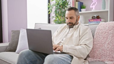 Sakallı, gri saçlı, evinde, kanepede dizüstü bilgisayarda çalışan olgun İspanyol bir adam..