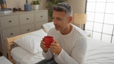 Yatak odasında elinde kırmızı bardakla oturan yakışıklı orta yaşlı bir adam, ev dekoru ve hafif ışıklandırmayla çevrili bir yatakta oturuyor, evinde bir anlık rahatlama yakalıyor..