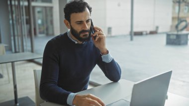 Orta yaşlı, sakallı bir adam telefonda bilgisayarla modern ofiste konuşuyor.