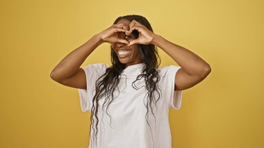 Kıvırcık saçlı neşeli Afrikalı kadın, sarı izole edilmiş arka plana kalp hareketi yapıyor.