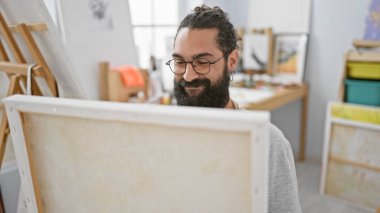 Parlak bir sanat stüdyosunda gözlüklü, gülümseyen sakallı adam yaratıcılık ve konsantrasyon yayıyor..