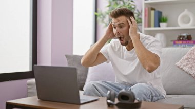 Oturma odasında sakallı, evdeki dizüstü bilgisayara tepki veren genç, şoke olmuş İspanyol bir adam..
