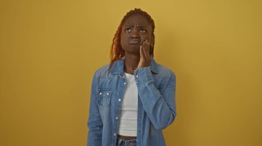 Kot giyen Afrikalı Amerikalı genç bir kadın parlak sarı bir arka plandan endişeli görünüyor..