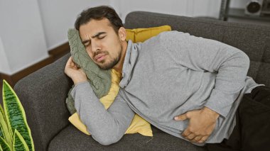 Sakallı genç bir İspanyol adam, kapalı bir kanepede dinlenirken rahatsız ya da rahatsız görünüyor, karnını sıkıyor..