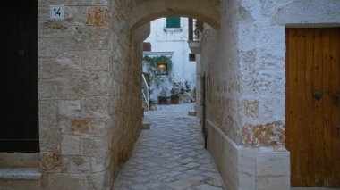 An empty narrow street in the old town of polignano a mare, italy, with stone walls leading to a quaint courtyard featuring green plants and warm lighting. clipart