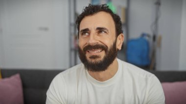 Oturma odasında gülümseyen sakallı bir adam rahatlamış yetişkin havası yayıyor..